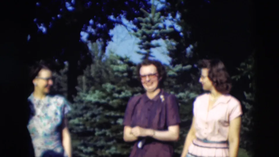 1951 Paducah home-movie frame showing three women in period dresses standing in a sunny garden near evergreen trees.