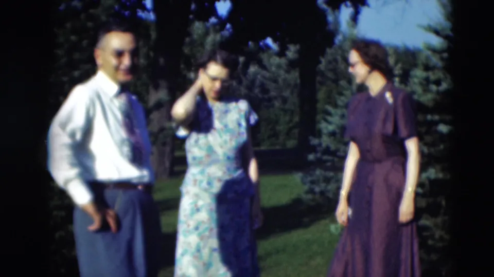 1951 Paducah frame with a man in a light shirt and tie standing near two women in dresses beside evergreen trees.