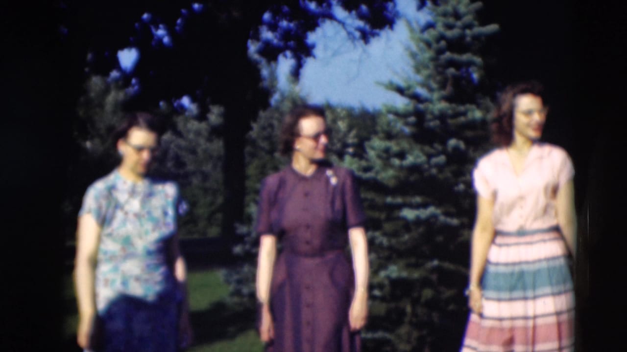 Three women in floral, purple, and striped dresses stand in a shaded Kentucky garden, facing different directions as they talk.
