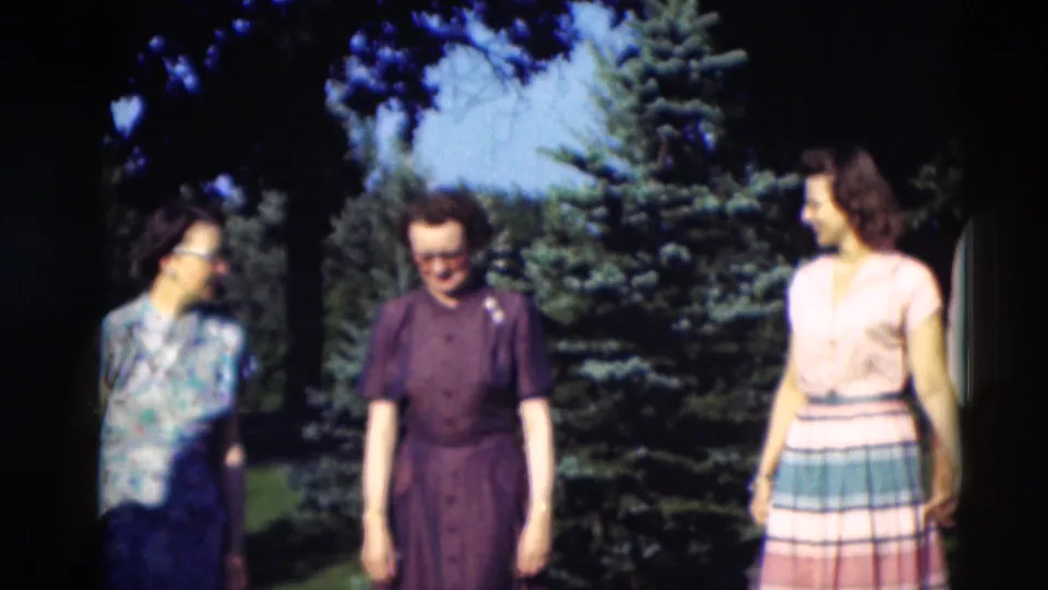 Three women in 1950s dresses appear to smile and converse in a Paducah garden, framed by shade, lawn, and evergreen trees.