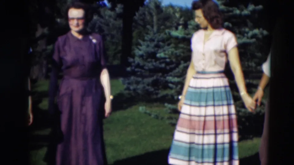 1951 Paducah frame with a woman in a purple dress standing near evergreens as another woman in a striped skirt turns toward a companion.