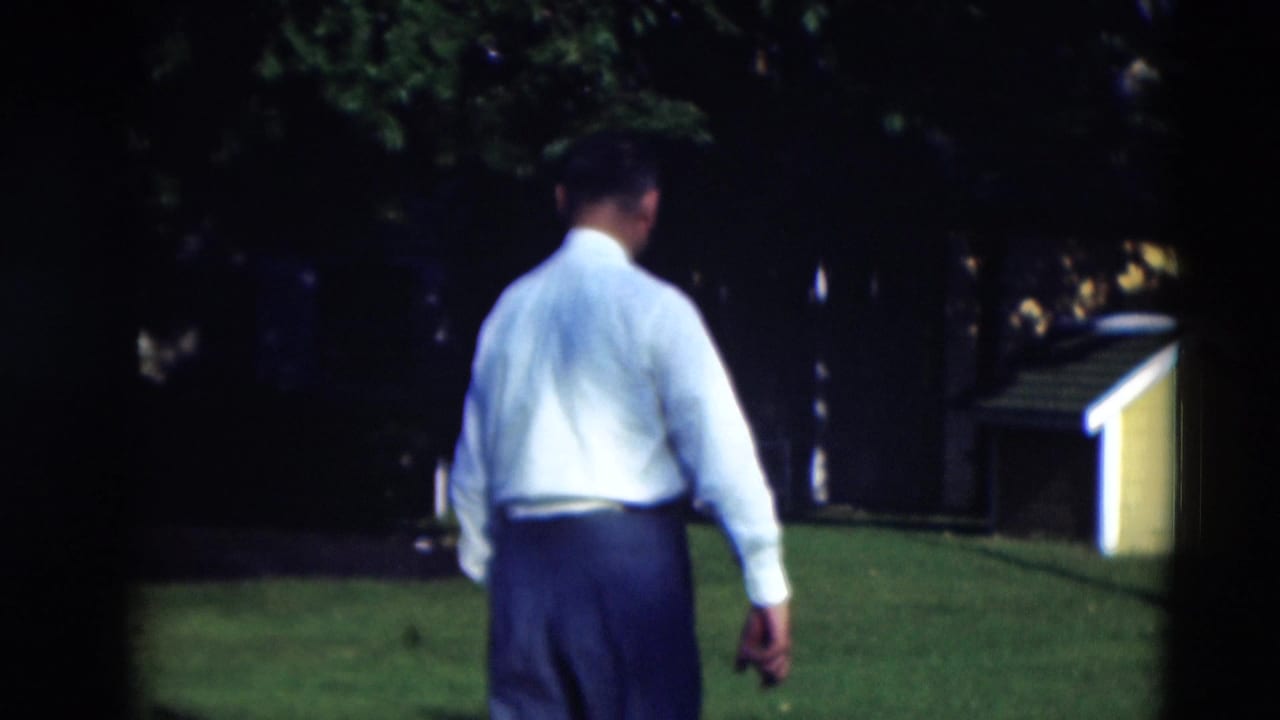 The man continues across the manicured lawn, framed from behind with trees and a small shed area in the background.
