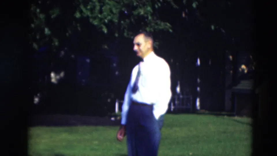 The man slows or pauses in side view, with his white shirt, dark tie, trousers, lawn, trees, and small yellow shed visible.