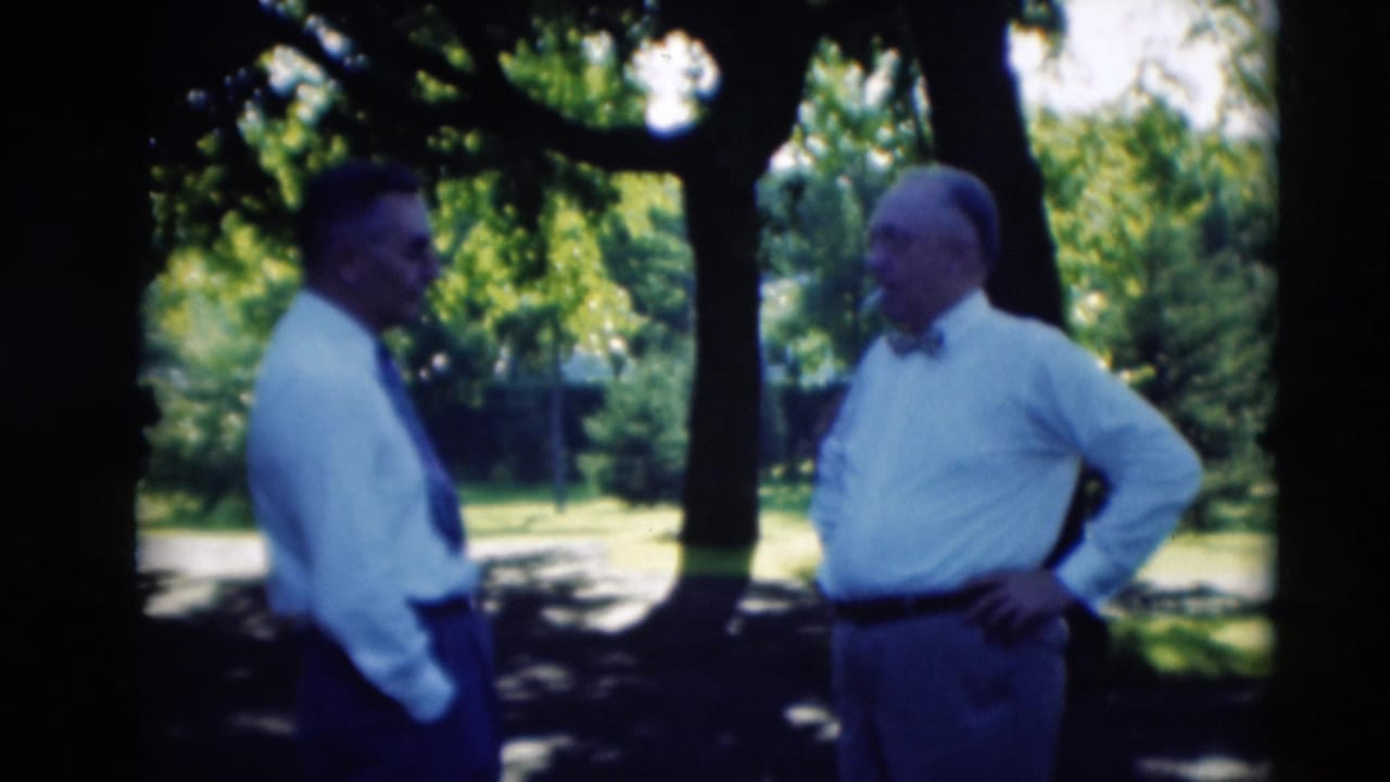 Two adult men in shirts and ties stand in conversation beneath trees, with sunlit grass behind them in Paducah, Kentucky.