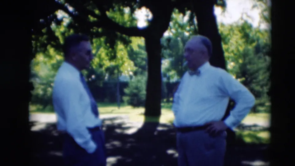 Two adult men in shirts and ties stand in conversation beneath trees, with sunlit grass behind them in Paducah, Kentucky.
