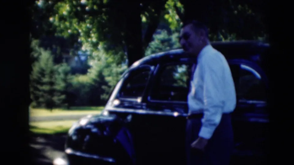 A man in a white shirt stands beside a dark rounded car on a lawn, with another figure moving through the shaded foreground.