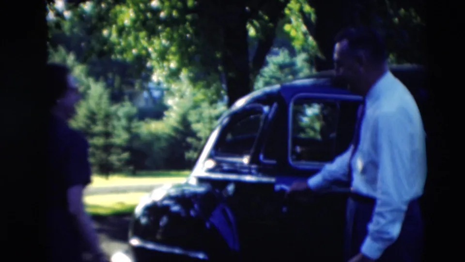 A man stands near the side door of a dark mid-century car under trees during an outdoor gathering in Paducah, Kentucky.