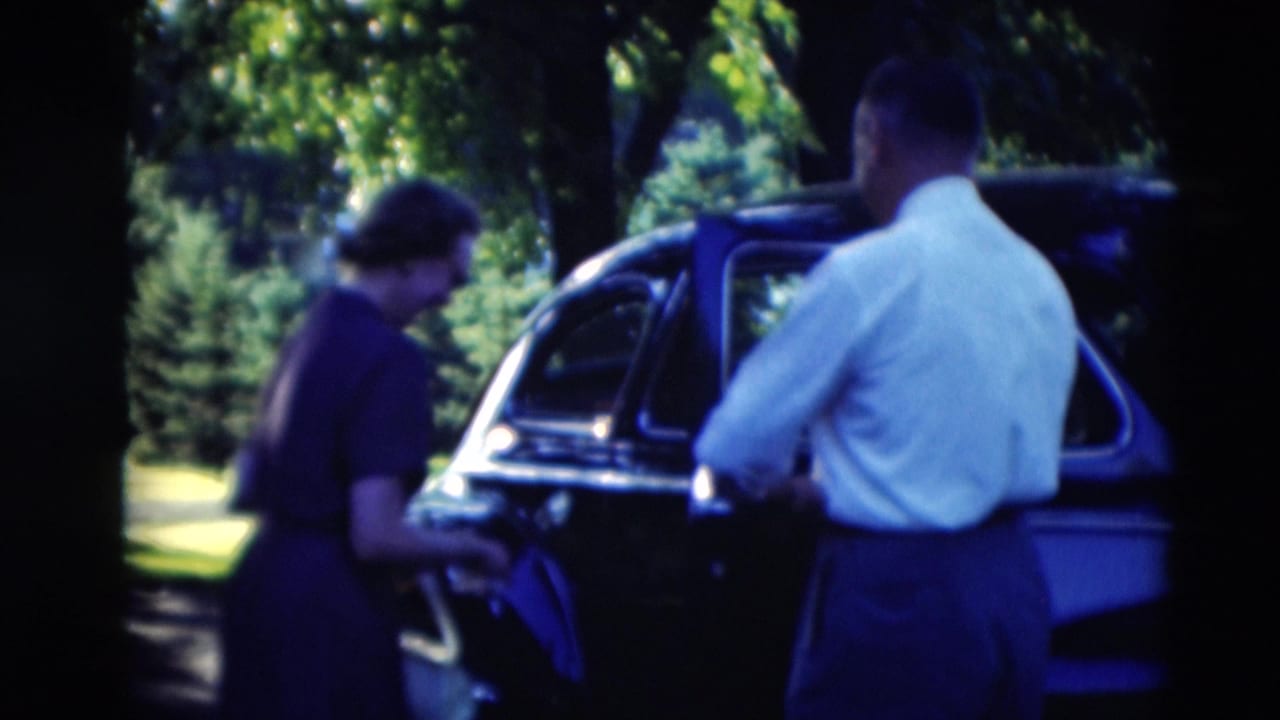 Man in a light shirt opens a dark vintage car door while a woman in a dark dress stands nearby.