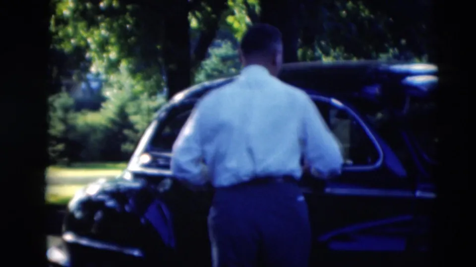 A man in a light shirt stands close to the open door of a dark car, with trees and lawn visible in the 1951 Paducah footage.