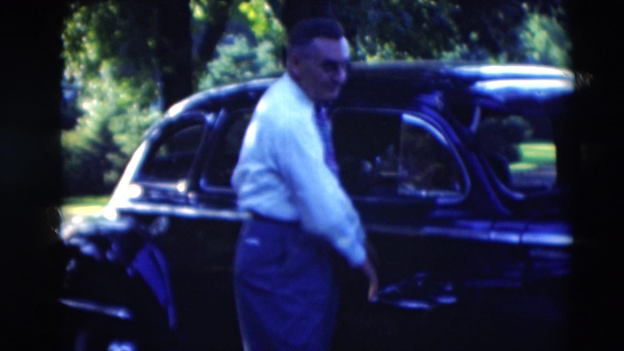 Man in a white shirt and tie stands beside a dark rounded car outdoors.