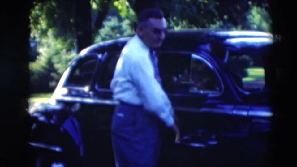 In Paducah, Kentucky, 1951, a man in a white shirt and tie stands by a black mid-century car in a leafy setting.