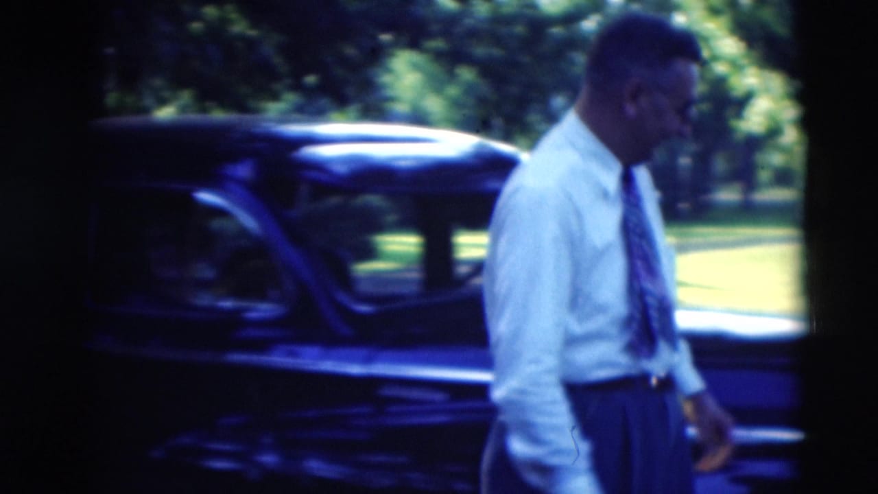 The man walks along the side of a black car, with trees and sunlit grass behind him in the 1951 Kentucky footage.
