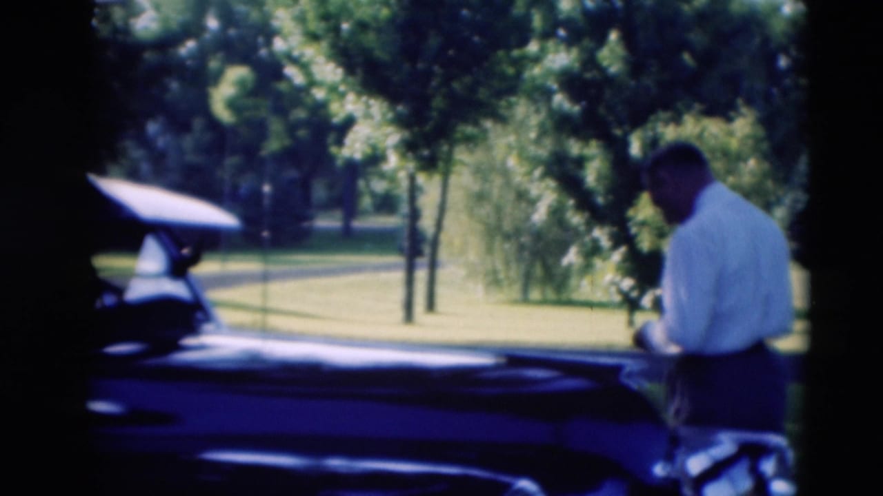 1951 Paducah home-movie frame showing a man beside a dark mid-century car with an open door on a green park lawn.