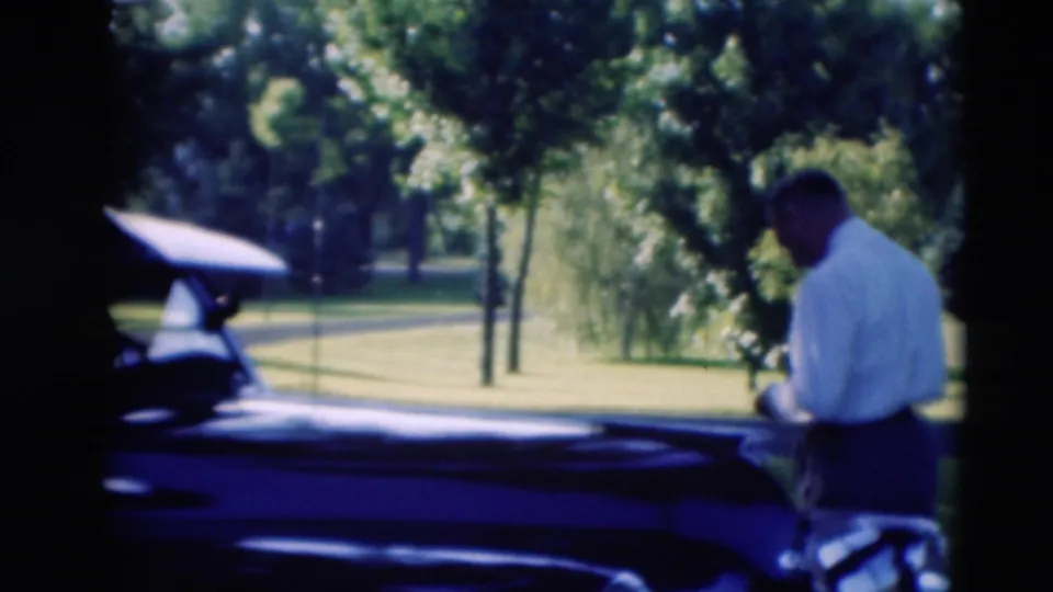 1951 Paducah home-movie frame showing a man beside a dark mid-century car with an open door on a green park lawn.