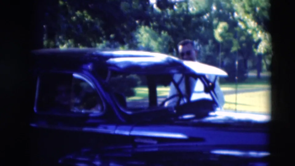 The dark mid-century car is seen in profile as the driver door remains open and the man is partly obscured behind it.