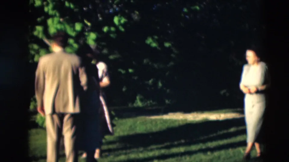 1951 Paducah home-movie frame showing adults gathered on grass as a woman in a light dress walks toward them from the right.