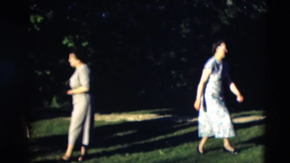Two adult women cross a green lawn in a 1951 Paducah home-movie scene with deep shade and foliage behind them.