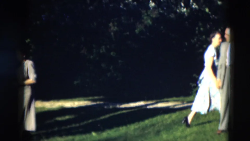 A woman in a patterned dress walks near a suited man on a shaded lawn in 1951 Paducah, Kentucky footage.