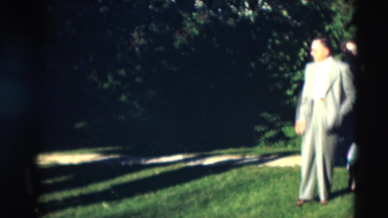 Man in a light suit standing on a sunlit lawn near trees.