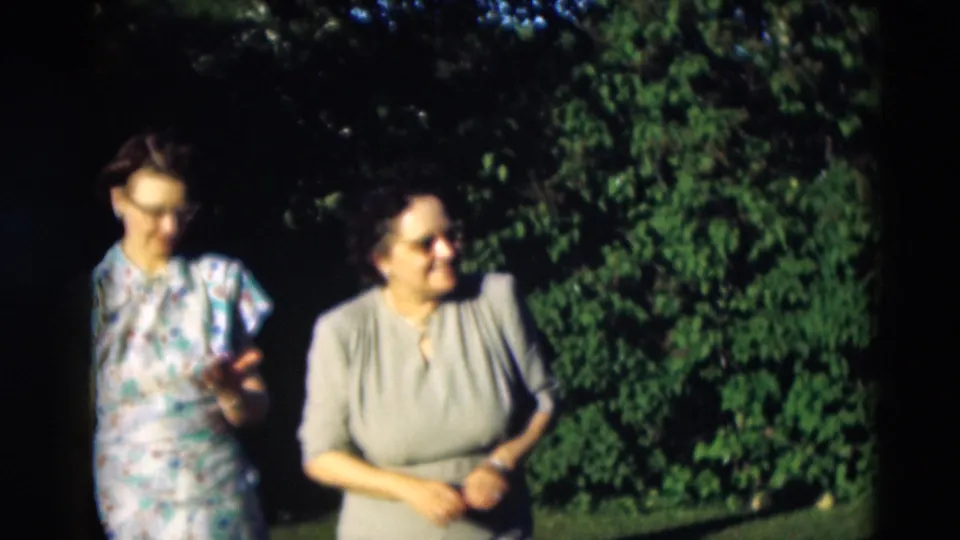 The two women continue together outdoors, with the woman in the blue floral dress gesturing as they move near trees.