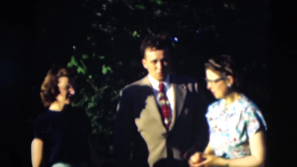 A young suited man walks outdoors with two women, including the woman in the blue floral dress, in the 1951 Paducah home movie.