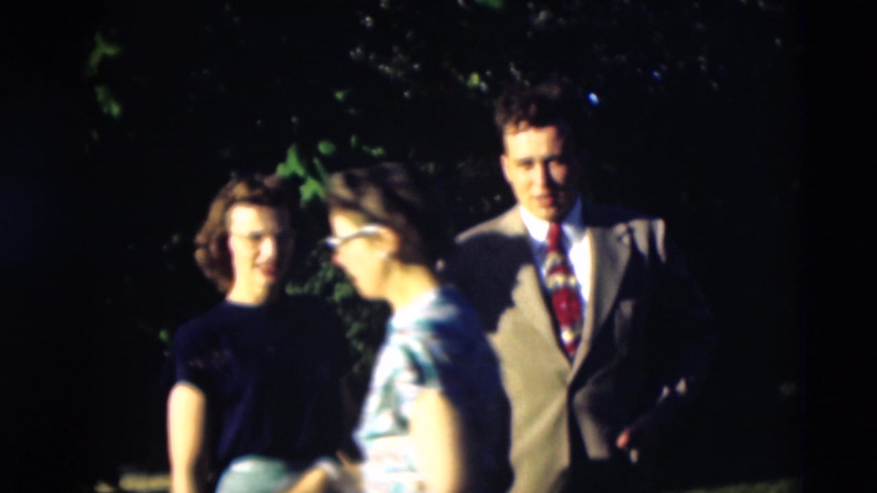 Three adults standing outdoors near leafy greenery, with a man in a light suit and two women nearby.