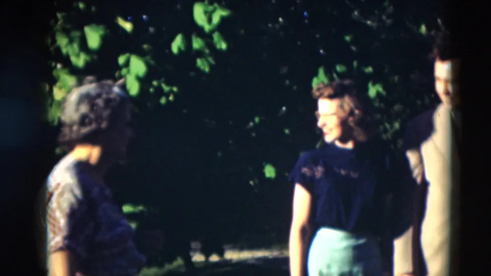 1951 Paducah home-movie frame showing adults shifting positions in a leafy garden, including a floral-dressed woman and a woman in a dark top.