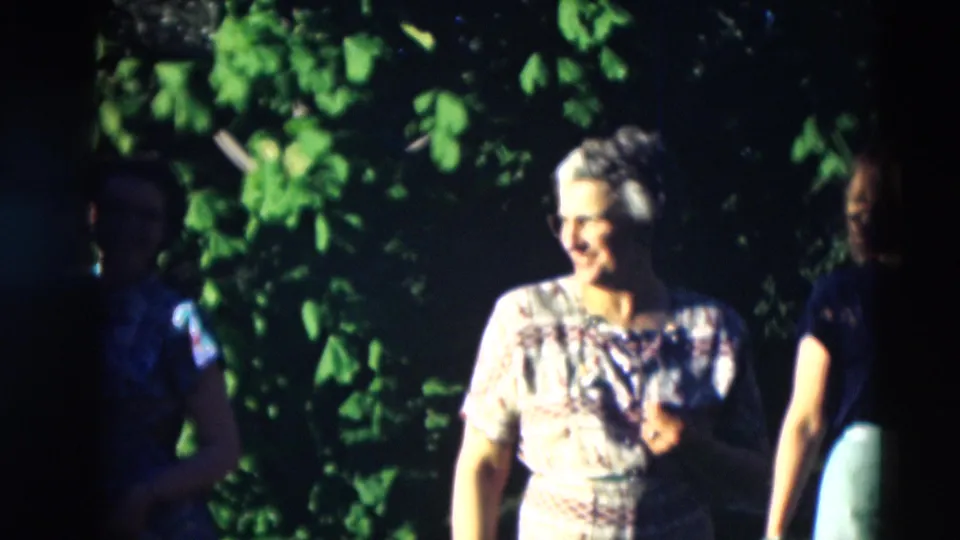 1951 Paducah garden frame with a smiling woman in a floral dress lit by sun, another woman partly visible, and leafy greenery behind them.