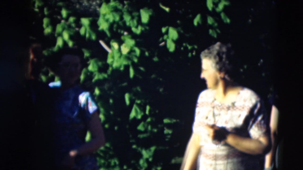1951 Paducah home-movie frame showing adults in a leafy garden, with one woman in patterned dress turned toward others.