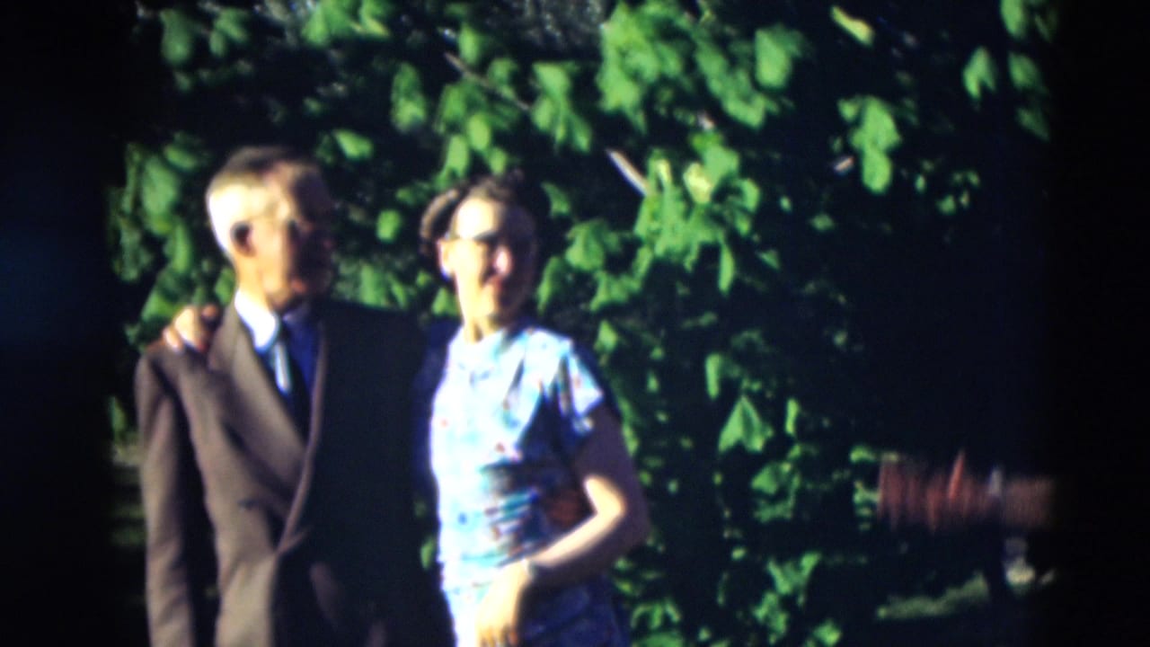 1951 Paducah frame of a suited man and a woman in a light dress standing arm in arm in a shaded garden.