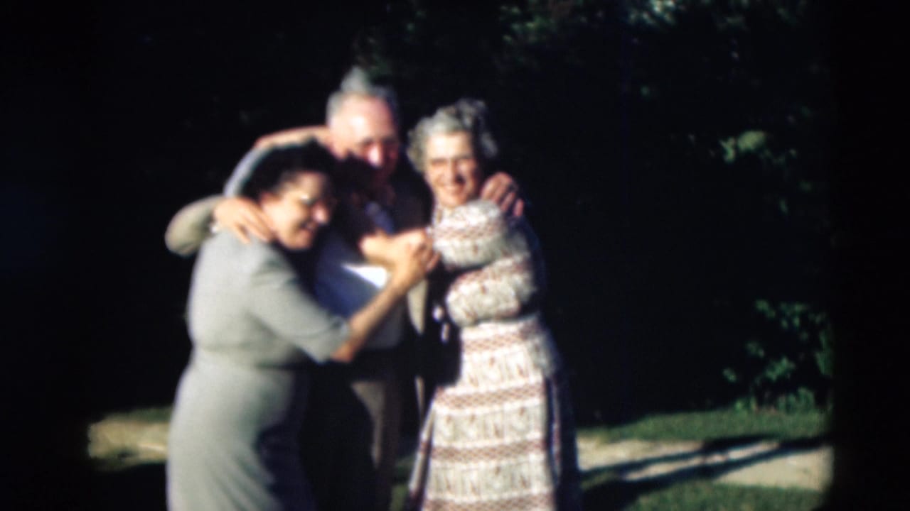 Three adults outdoors lean together in a smiling group embrace.