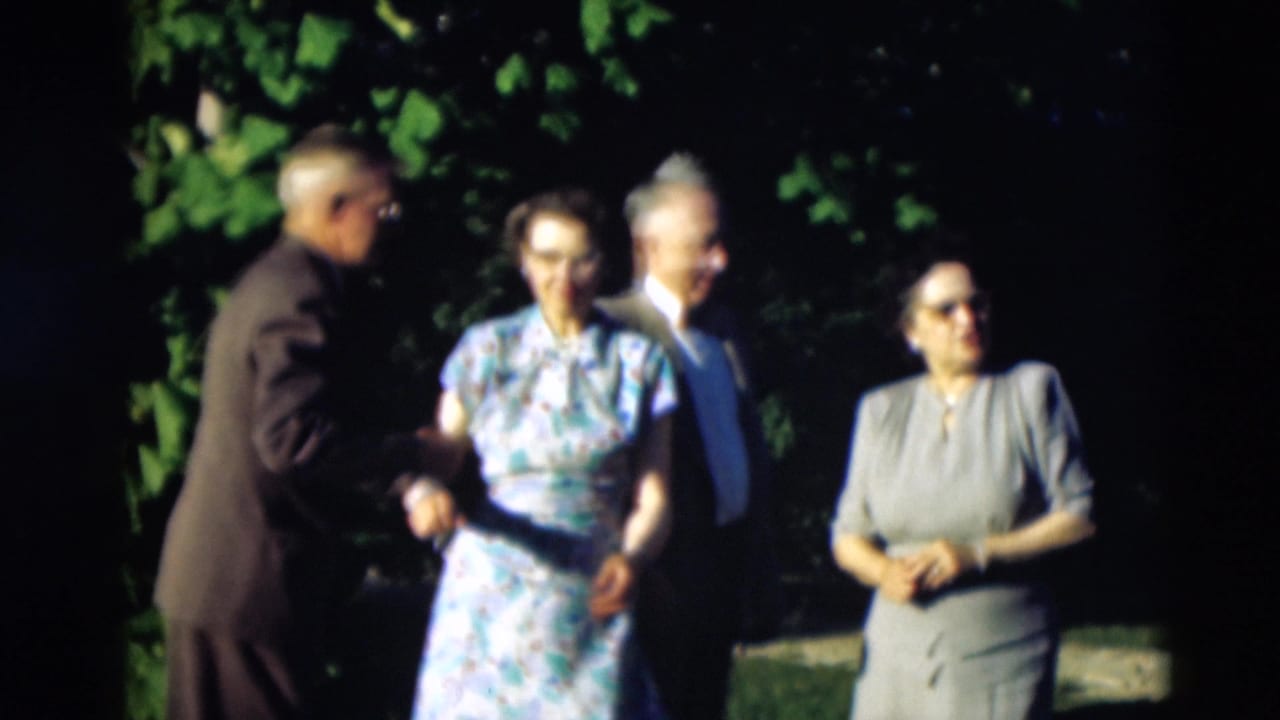 Four adults stand outdoors as one man reaches toward a woman in a floral dress.
