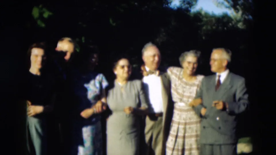 The group faces forward in a shaded garden area, with adults in 1950s formal clothing linked together for a photograph.