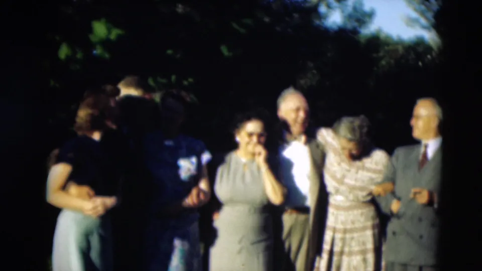 1951 home-movie frame of adults gathered under trees, including a woman with eyeglasses and others standing arm in arm.