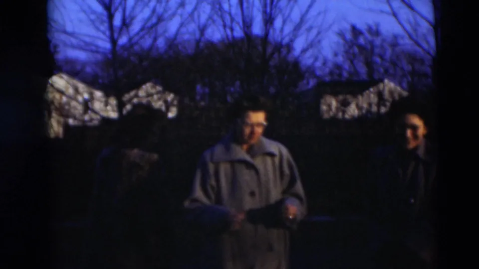 Adults in coats stand in a dim outdoor yard at twilight, with bare trees and house roofs visible in the background.