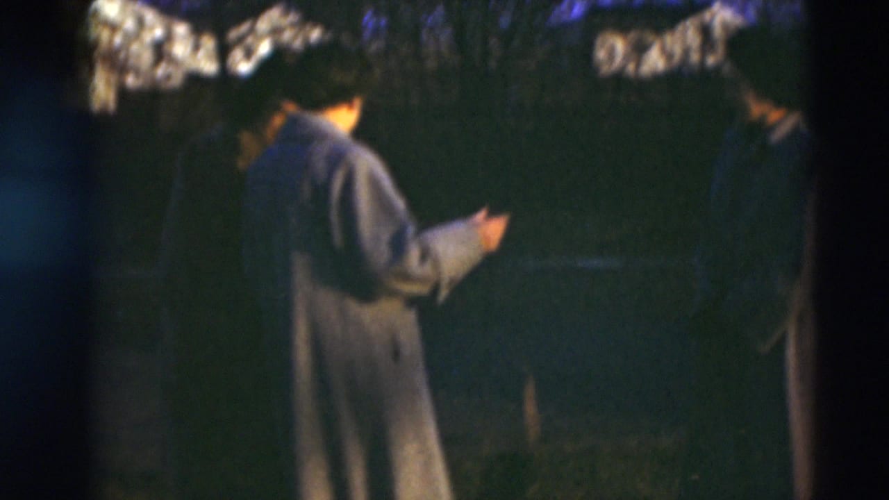 Two women in long coats stand in profile outdoors in the 1951 Paducah home-movie footage, with dark trees and houses in the background.