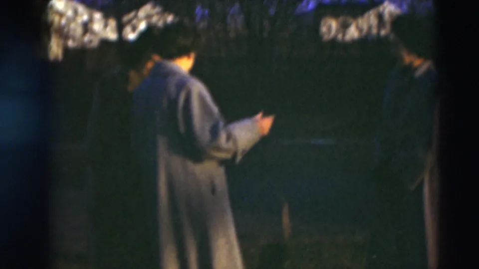 Two women in long coats stand in profile outdoors in the 1951 Paducah home-movie footage, with dark trees and houses in the background.