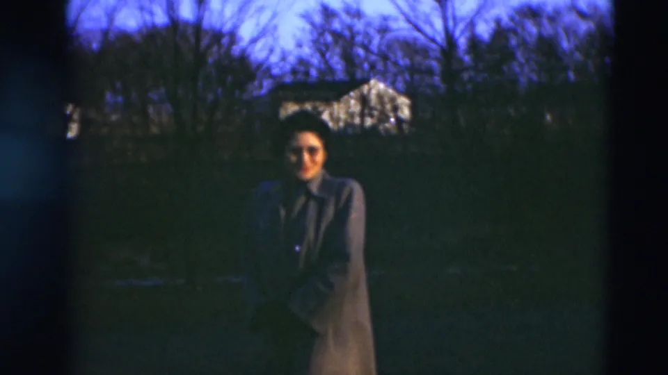 A woman in a long coat smiles or looks toward the camera in the 1951 Paducah outdoor home-movie scene, with trees and a house behind her.
