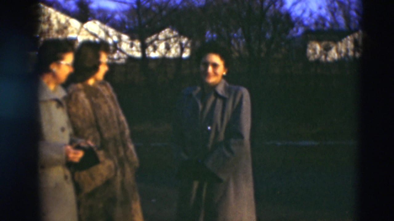 Three adults in long coats gather in a Paducah yard at dusk, with bare trees and distant houses behind them in 1951 8mm footage.
