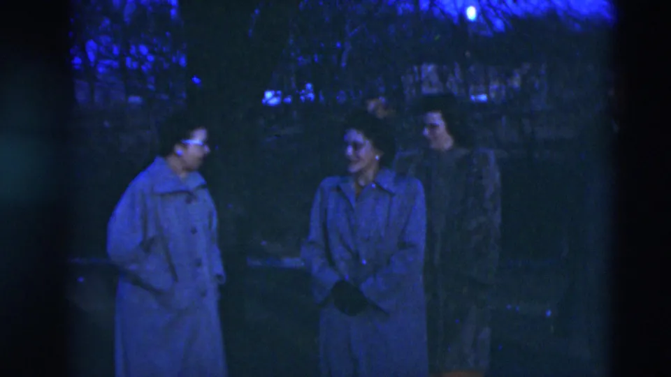 Two women in long coats appear to converse in a dim Paducah yard, with a third adult partly visible behind them in 1951 home-movie footage.