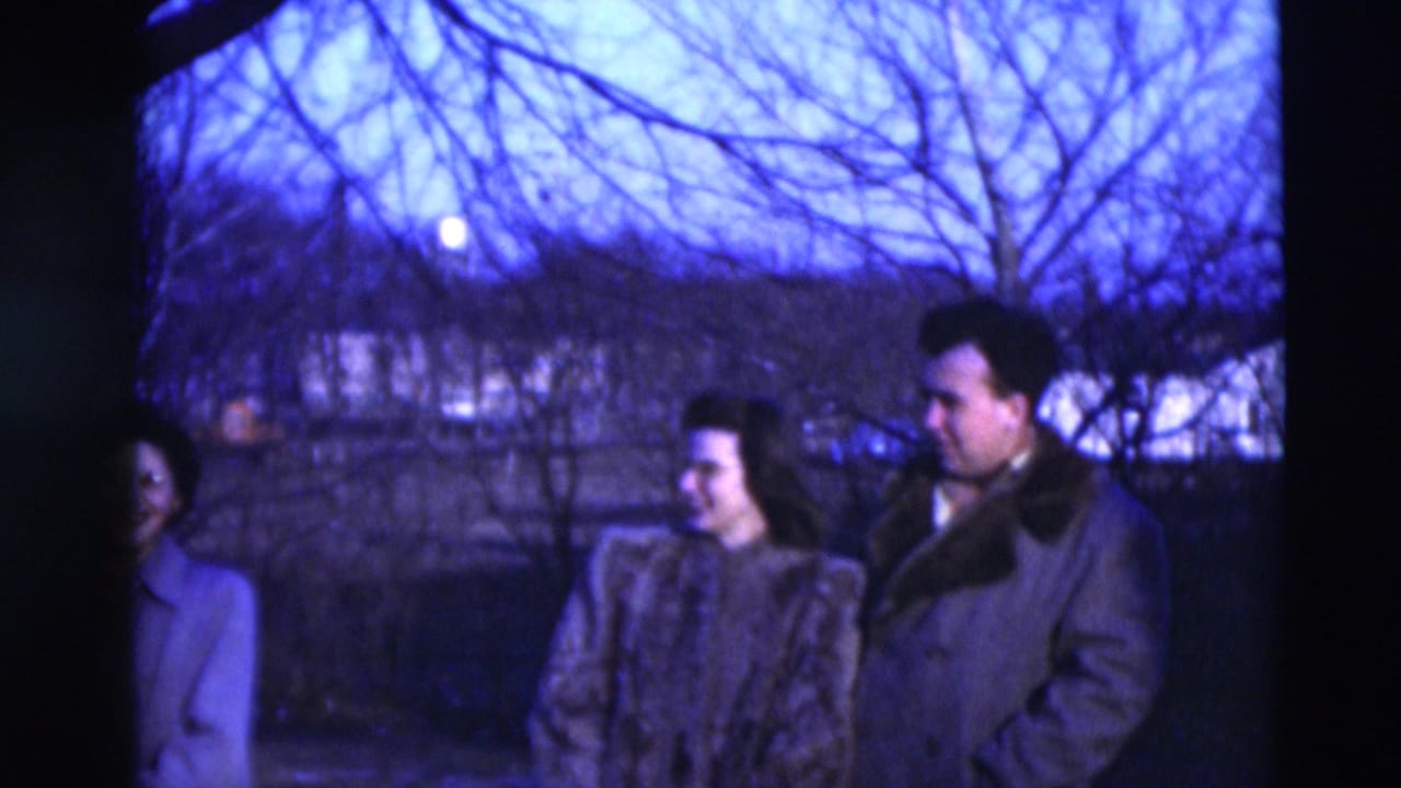 Three adults in 1951 Paducah home-movie footage stand near a dark tree trunk, dressed in winter coats under a blue evening sky.