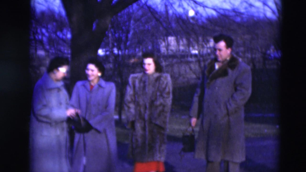Four adults in winter coats stand near bare trees on a residential street.