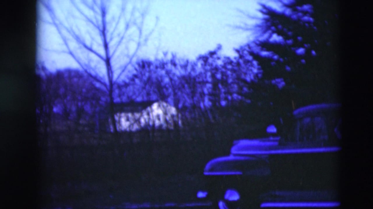 Blue-purple 8mm frame showing a parked rounded car beside trees and a distant house.