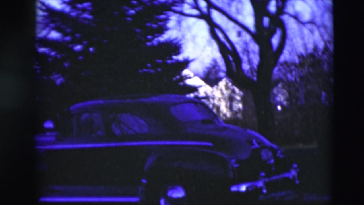 1951 Paducah home-movie frame showing an older dark car near trees, with strong purple-blue tint and soft 8mm texture.