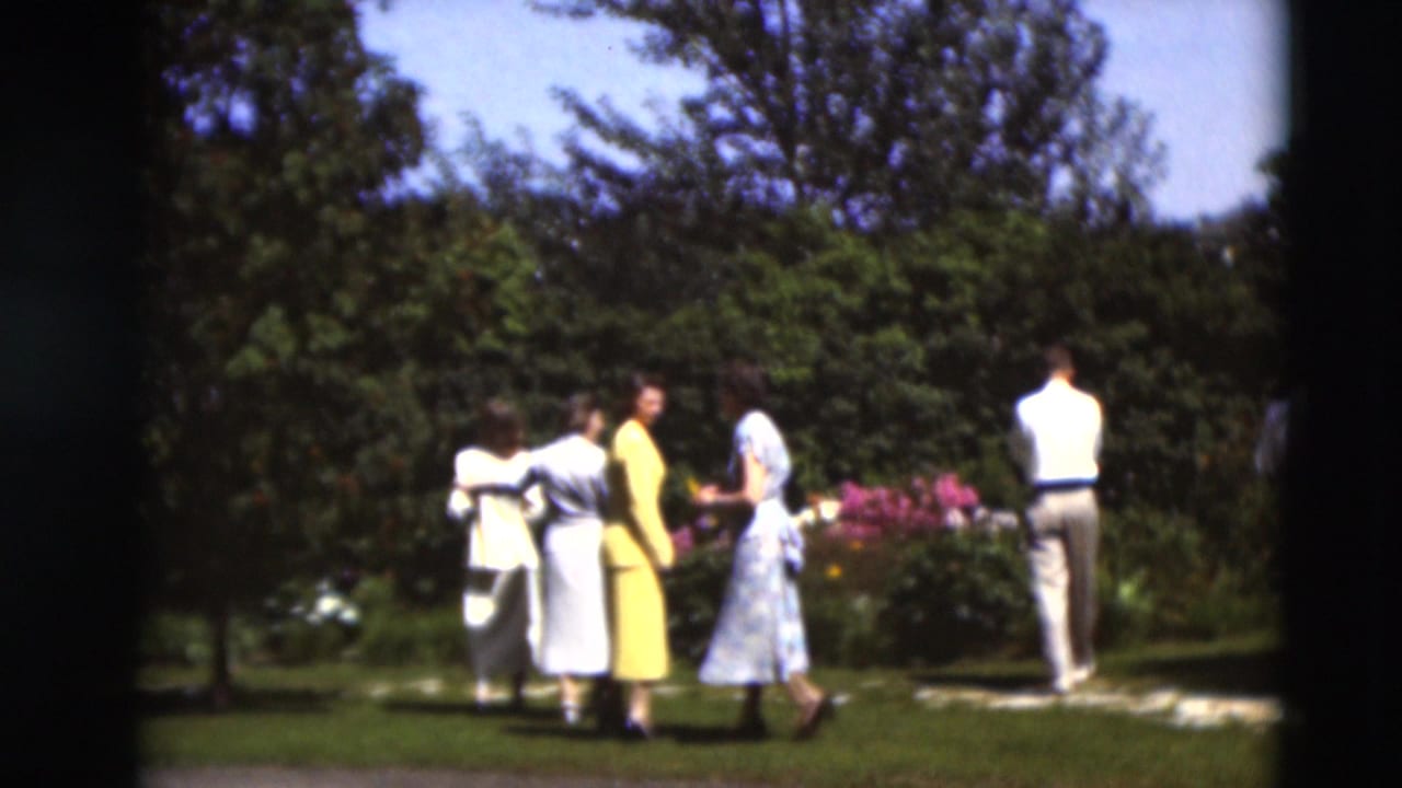 Adults stand and walk near flower beds and shrubs in a sunny garden.