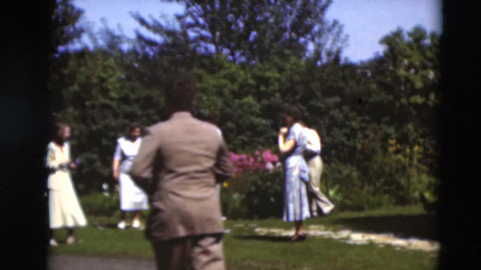 A man in a suit seen from behind approaches adults near flower beds in 1951 Paducah, Kentucky garden footage.