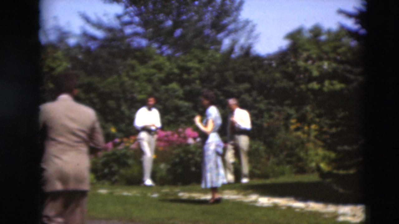 Adults in dress clothes stand and walk near a garden path and flowering beds.