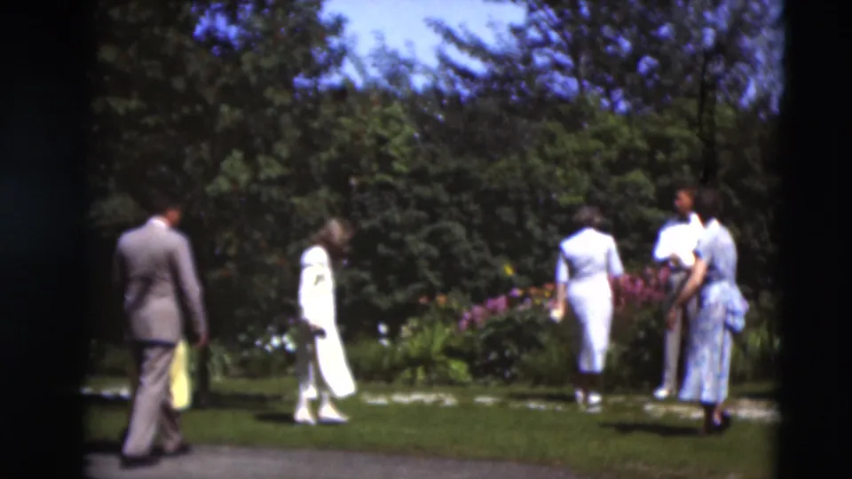 Several adults in 1951 Paducah home-movie footage walk through a sunlit garden bordered by flowers and trees.