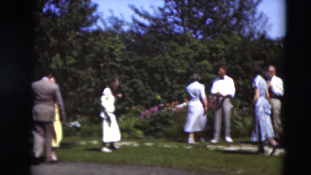 1951 Paducah home-movie frame showing adults in mid-century clothing gathered beside a grassy path and blooming garden beds.
