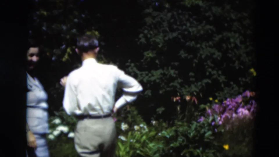 In 1951 Paducah home-movie footage, adults stand in a garden with purple blooms, green foliage, and bright sunlight filtering through shade.